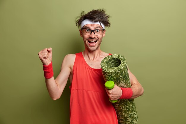 Cheerful positive man works out with dumbbell, raises arm and clenches fist, carries karemat, has aim to have fitness muscular body feels powerful and strong dressed in red shirt, headband, wristbands