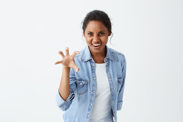 Cheerful positive playful having fun dark-skinned woman with dark hair dressed in light-blue denim shirt, showing her teeth and actively gesturing, trying to scare someone.