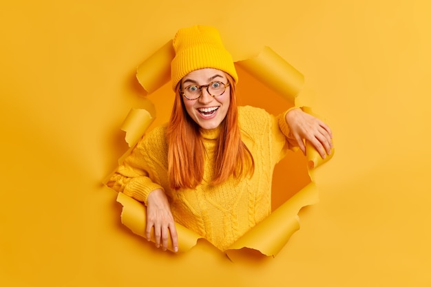 쾌활한 긍정적 인 빨간 머리 여자는 기쁜 표정으로 보이는 좋은 분위기가 찢어진 종이를 통해 사진을 위해 포즈를 취하는 것을 기쁘게 생각하는 노란 모자와 스웨터를 입습니다.