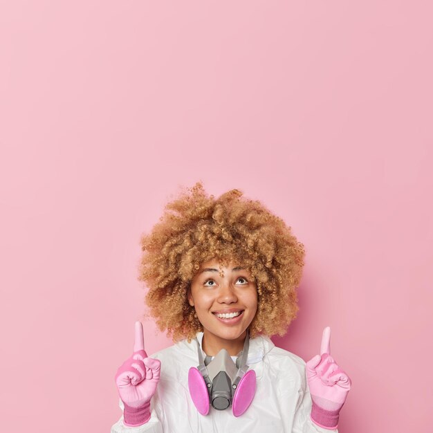 Free photo cheerful positive woman dressed in chemical suit and gloves points overhead warns you about environmental hazard has glad smile on face isolated over pink background blank space for your text