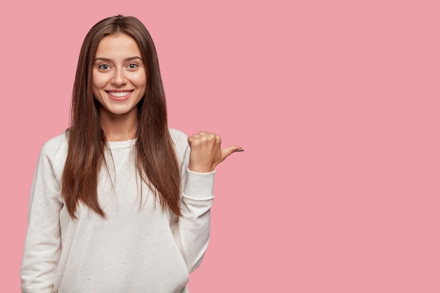 Cheerful positive young European woman with dark hair, broad shining smile, points with thumb aside