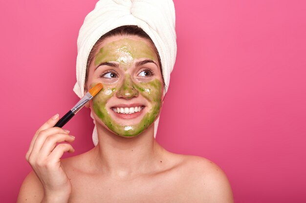 Cheerful positive young female putting colorful mask on her face with help of professional brush, looking aside, being happy. Attractive girl enjoys spending time doing beauty procedures for skin.