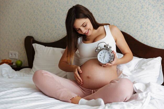 Cheerful pregnant woman sitting on bed holding alarm