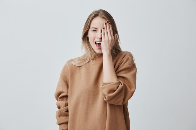 Cheerful pretty charming woman in loose sweater with fair hair smiling happily, having fun indoors, closing one eye with hand.