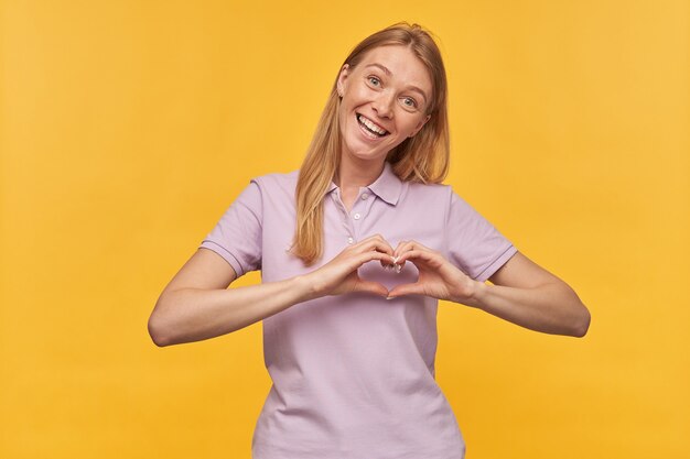 Cheerful pretty woman with freckles in lavender tshirt showing heart shape by hands and smiling on yellow