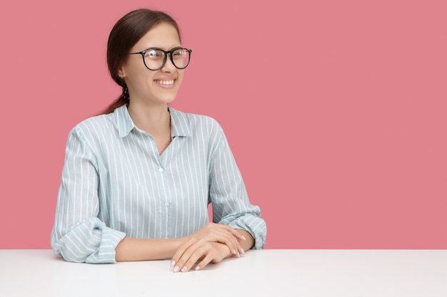 Cheerful pretty young female manager in glasses laughing at joke or funny situation, having fun at work, sitting at desk. Positive human facial expressions, emotions, reaction and feelings