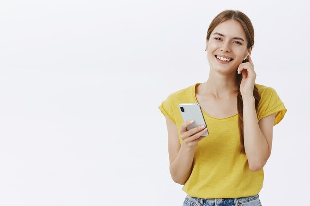 Cheerful pretty young girl listening music in wireless earphones, holding smartphone, enjoying podcast