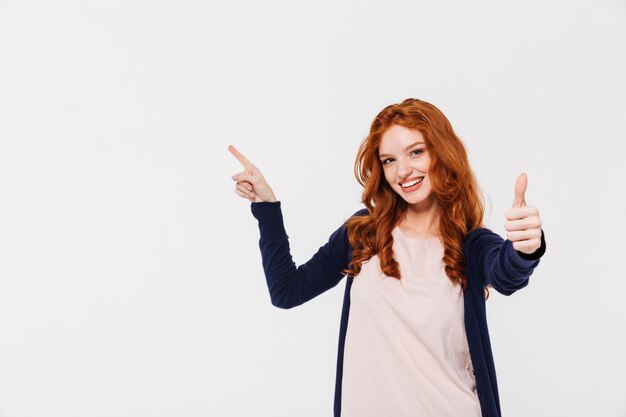Cheerful pretty young redhead lady showing thumbs up while pointing.