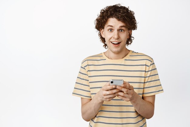 Cheerful queer guy holding smartphone looking excited at camera standing in tshirt over white background