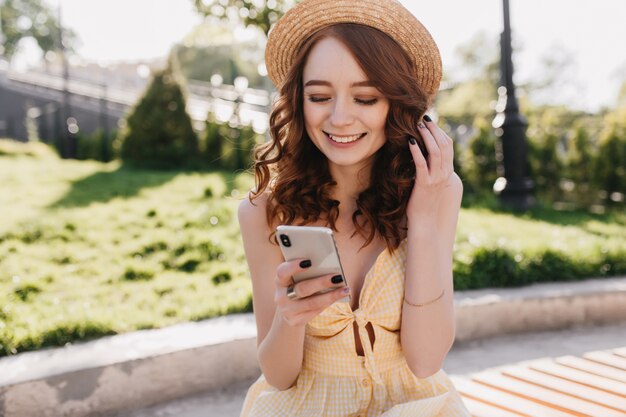 Cheerful red-haired girl in cute hat read messages with happy smile. Outdoor photo of lovable ginger woman in yellow dress relaxing in park in morning.