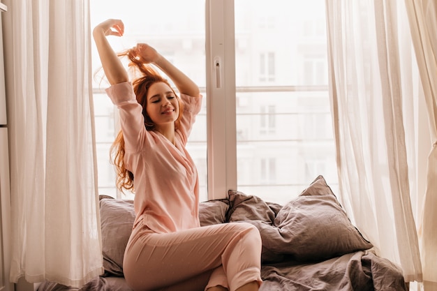 Cheerful red-haired girl in pajama smiling in morning. Merry ginger woman posing in bed.