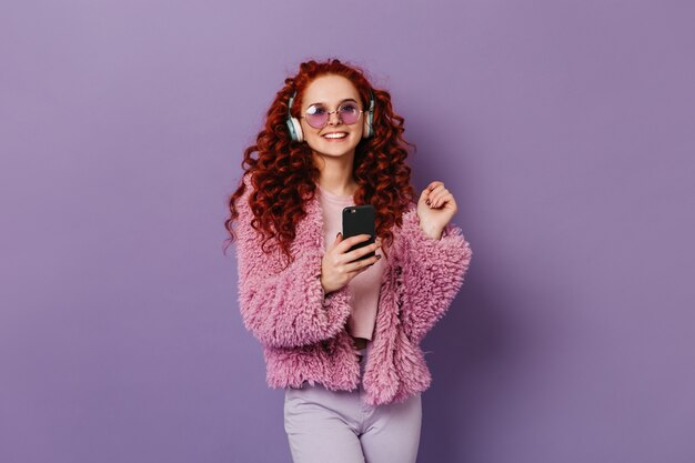 Cheerful red-haired girl in pink eco-coat and light pants smiling. Woman in blue headphones holds black smartphone.