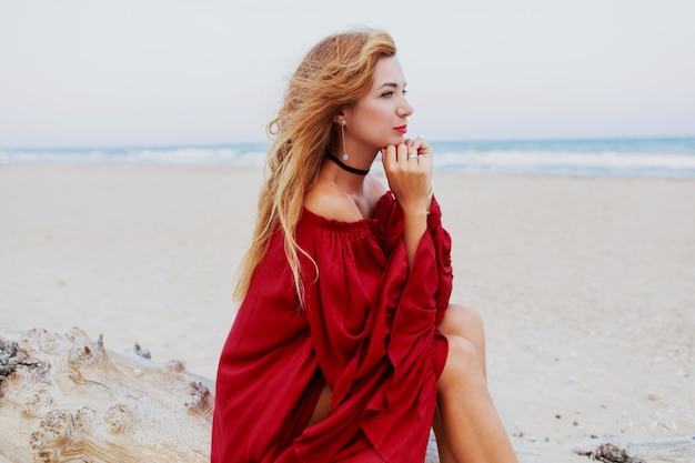 Cheerful red-head girl posing on beach. Sitting on white sand. Windy hairs. Trendy outfit. Lifestyle portrait. Travel mood. Ocean coast.