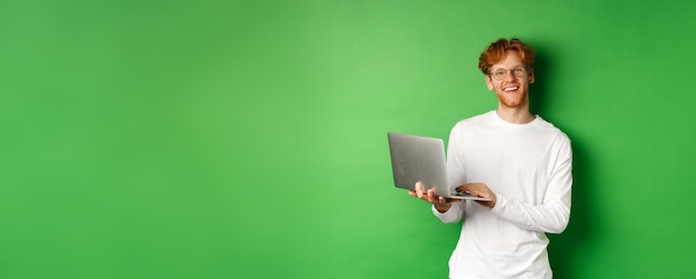Бесплатное фото Веселый рыжий фрилансер в очках улыбается в камеру, работающую на ноутбуке, стоя над зеленым
