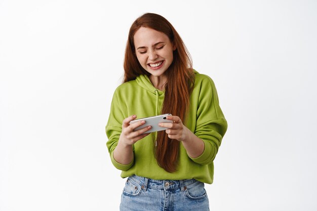 Cheerful redhead girl playing on video game mobile phone, watching funny video on smartphone and laughing, standing on white