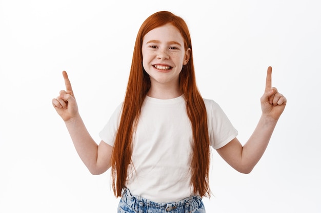 Бесплатное фото Веселая рыжая девушка улыбается с зубами и указывая пальцами вверх. рыжий ребенок с веснушками счастлив, показывая рекламу, белая стена