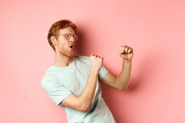 Cheerful redhead guy listening music and dancing triumphing or celebrating success standing over pin...