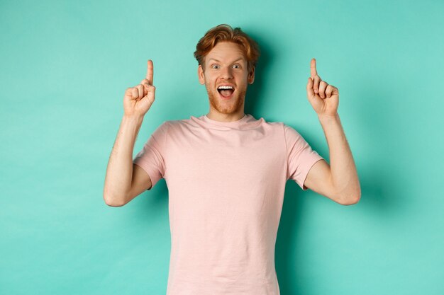 ターコイズブルーの背景の上に立って、指を上に向け、カメラを畏敬の念を持って見つめ、広告を表示しているTシャツの陽気な赤毛の男。