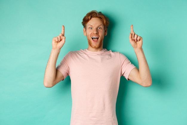 Веселый рыжий мужчина в футболке указывая пальцами вверх, с трепетом глядя в камеру и показывая рекламу, стоя на бирюзовом фоне.