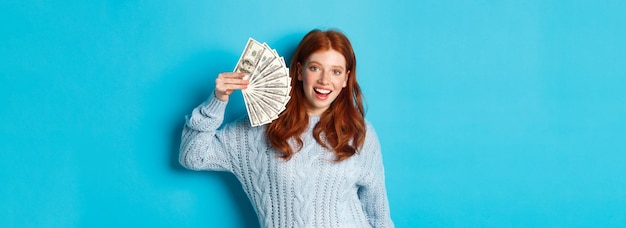 Foto gratuita donna allegra della testarossa in maglione che mostra i dollari che sorridono soddisfatti e che tengono i soldi che controllano il bl