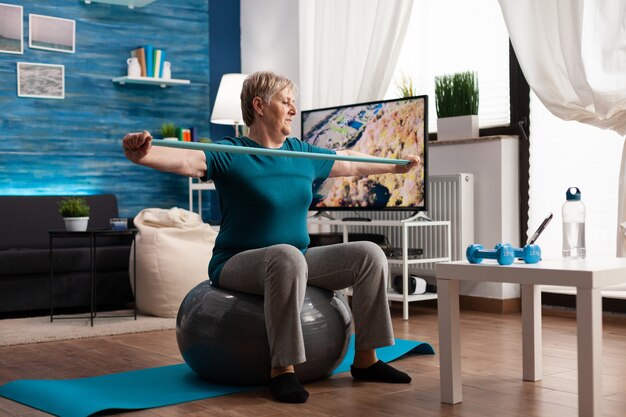 有酸素運動を練習するゴムバンドを使用して腕の筋肉を操作する陽気な引退した年金受給者。ジムのビデオを見ている体のヘルスケアの抵抗で働いている居間でスイスのボールに座っている年金受給者