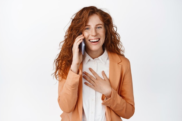 Cheerful saleswoman having phone call talking on smartphone and laughing smiling amused standing over white background