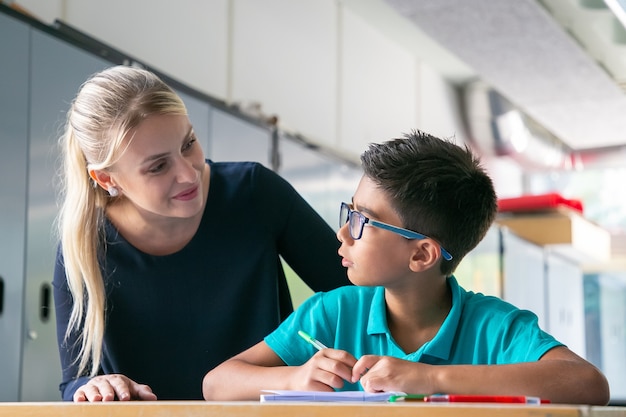 Cheerful school teacher giving help and support to schoolboy in class