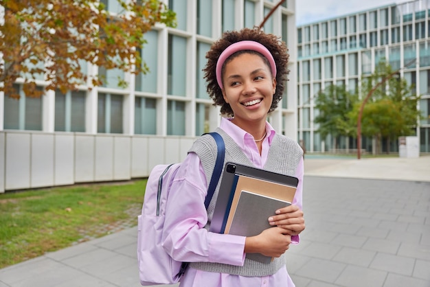 Cheerful schoolgirl carries rucksack notepads and digital tablet looks into distance with happy expression walks in campus during daytime Female student returns from university Education concept