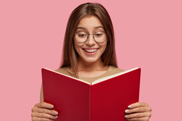 Cheerful schoolgirl holds textbook, reads interesting book