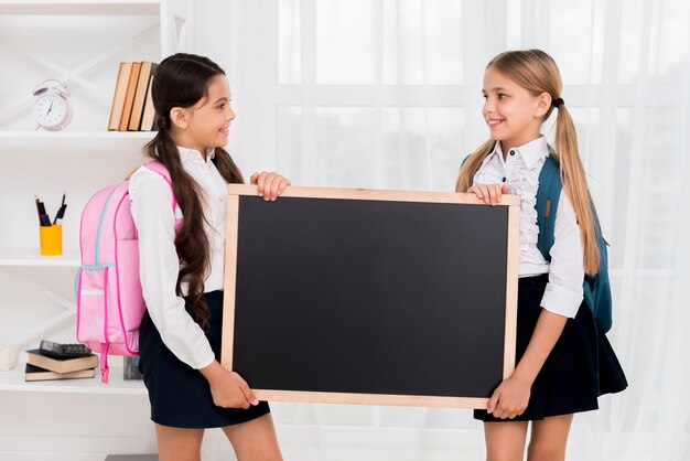 Cheerful schoolgirls with backpacks holding blackboard in room