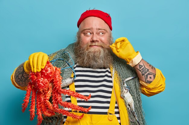 Cheerful seaman curls mustache, holds big sea creature, dressed in striped jumper and yellow overalls