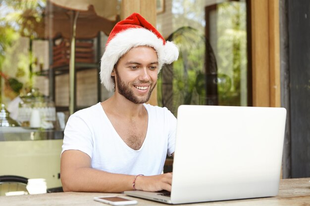 Cheerful self-employed male wearing Santa Claus hat working remotely on laptop pc, sitting at cafe table with cell phone, spending winter in hot country