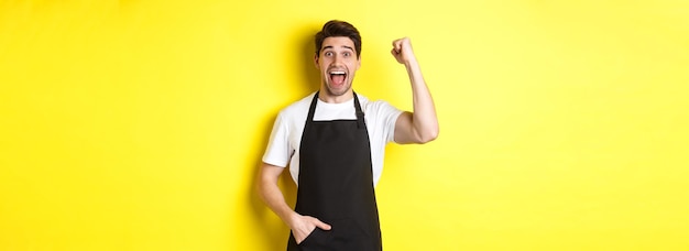 Cheerful seller making fist pump rejoicing and triumphing standing in black apron against yellow bac