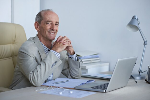 Cheerful senior Caucasian executive sitting at desk in office and smiling for camera