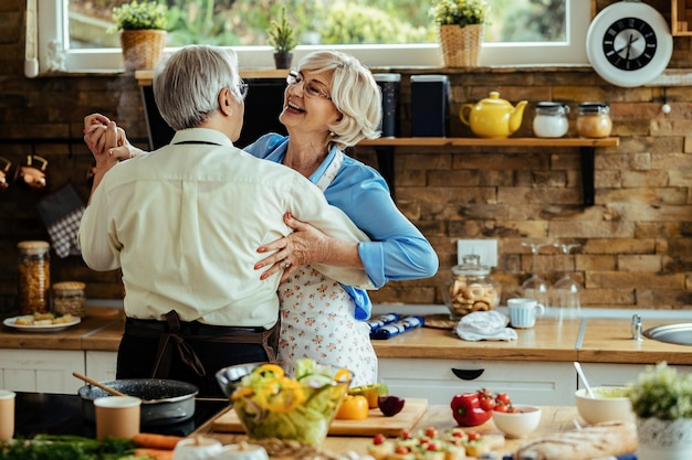 Cheerful senior couple having fun and dancing while preparing lunch in the kitchen.