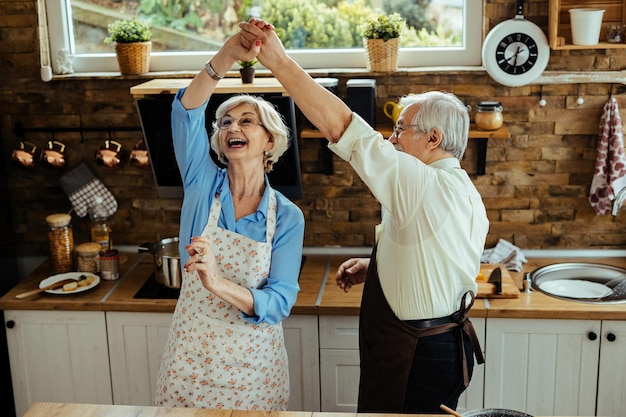 Cheerful senior husband and wife having fun while dancing in the kitchen.