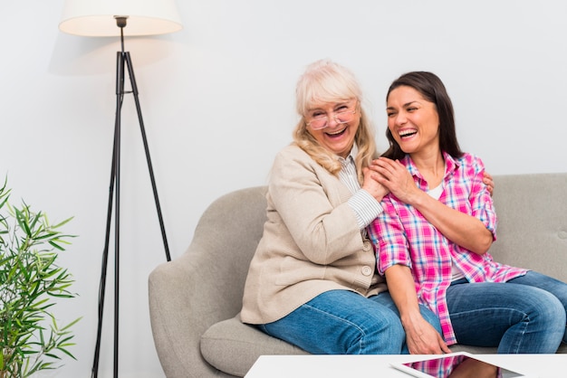 Free photo cheerful senior mother and her daughter sitting on sofa with digital tablet on white table