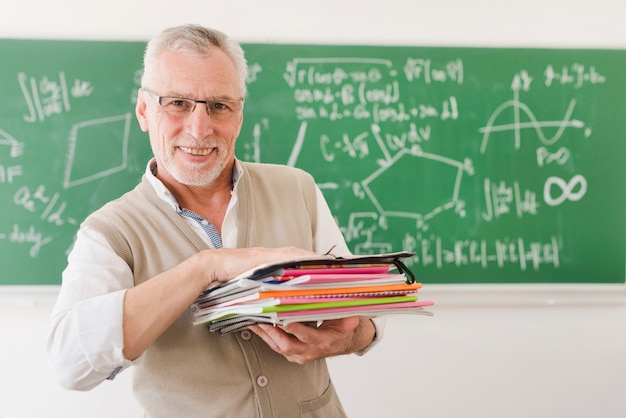 Cheerful senior professor holding pile of notebooks in lecture room