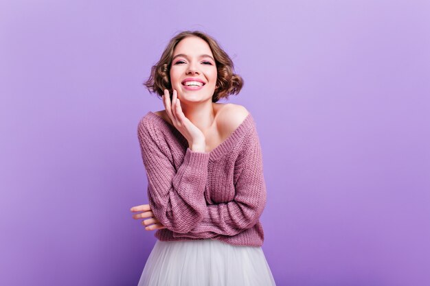 아름다운 스웨터 사진 촬영을 즐기는 쾌활한 짧은 머리 소녀. 보라색 벽에 웃 고 행복 로맨틱 화이트 레이디의 실내 초상화.