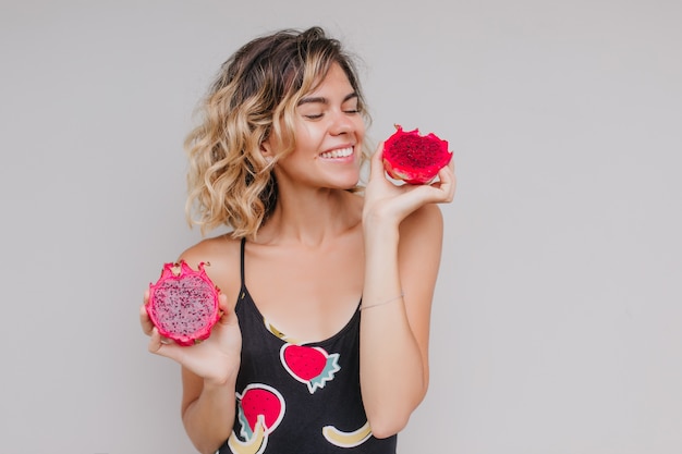 cheerful short-haired girl looking at juicy pitaya. Indoor photo of spectacular tanned woman isolated with dragon fruit.