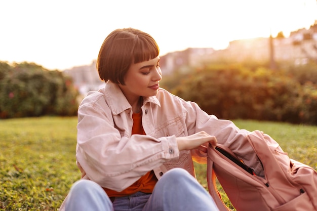 Cheerful shorthaired woman opens backpack outdoors Joyful young girl on pink denim jacket and jeans sits on grass