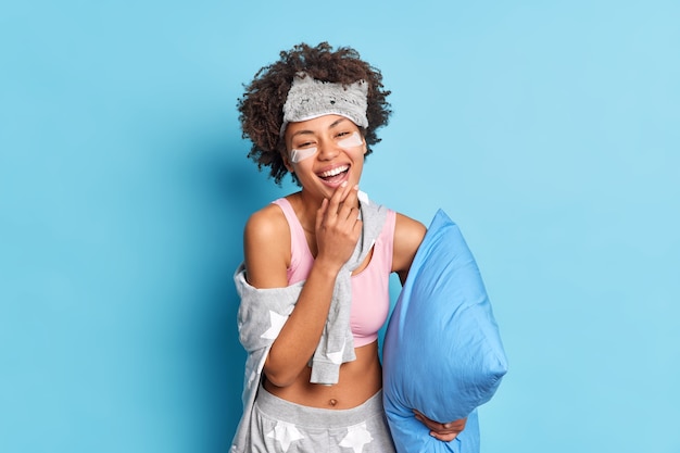 Cheerful sincere ethnic curly girl in nightwear smiles broadly prepares for sleep holds pillow has good mood isolated over blue background undergoes beauty treatments.