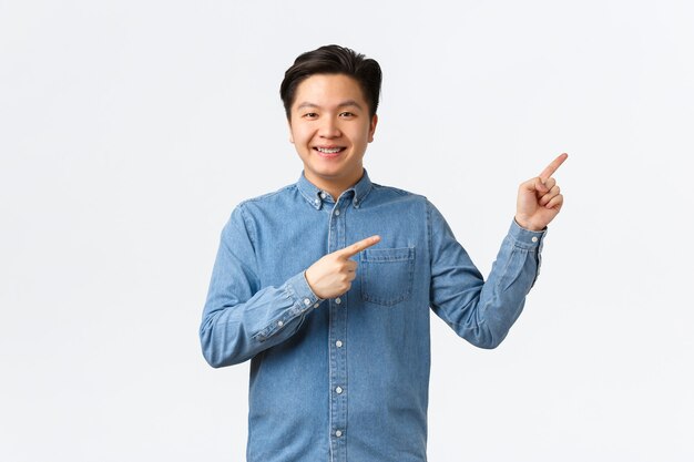 Cheerful smiling asian young man with braces in blue shirt, pointing fingers upper right corner proud and confident, showing link or make announcement, standing white background