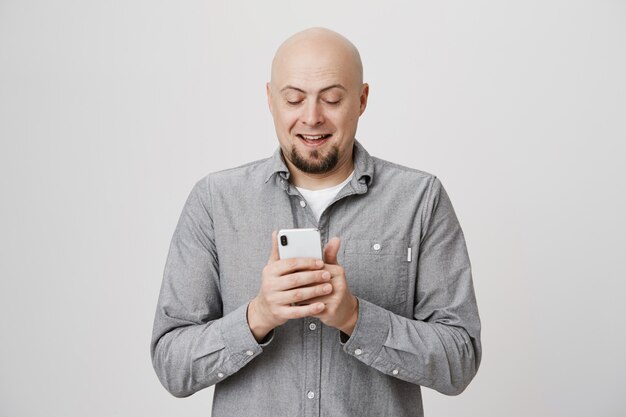 Cheerful smiling bald adult man messaging, using smartphone