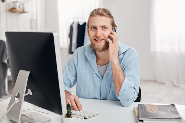 Cheerful smiling bearded male student recieves call from friend, sits at light office, dressed in blue shirt, finishes work soon. Handsome male freelancer has phone conversation, discusses ideas.