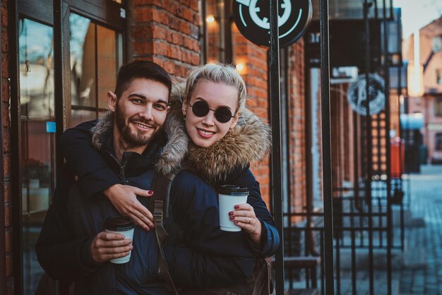 쾌활한 웃는 커플이 아늑한 카페테리아 밖에서 커피를 즐기고 있고, 여자가 남자를 껴안고 있습니다.