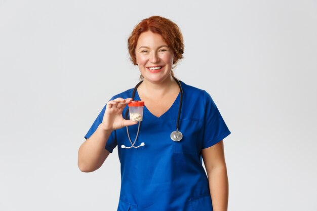 Cheerful smiling female meical worker, doctor in scrubs showing container with vitamins or medication, recommend pills, standing 