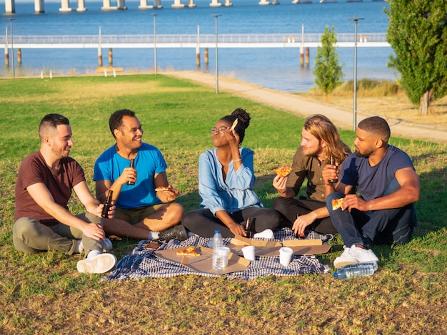 Веселые улыбающиеся друзья, пикник в парке. Молодые люди сидят на зеленой траве и едят пиццу. Концепция пикника