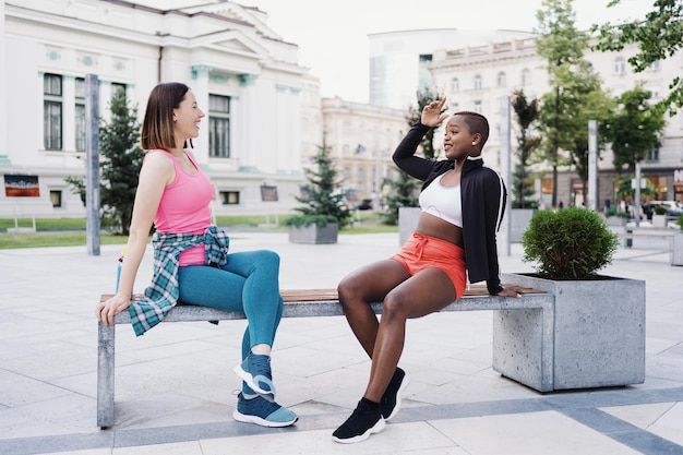 운동복을 입은 쾌활한 웃는 친구들은 도시의 벤치에 앉아 공원에서 토론하며 운동을 하는 다민족 여성