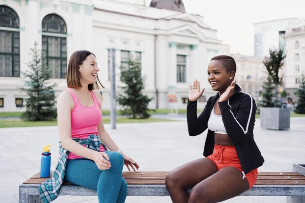 Cheerful smiling friends in sportswear sitting on bench in the city dicussing in park Multiethnic women having a fitness workout break